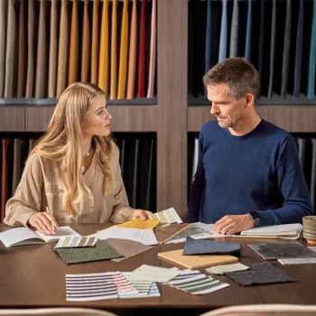A woman and a man sit at a table covered with fabric samples, swatches, and catalogs, discussing interior design options. Behind them, shelves display neatly arranged rolls of colorful fabric.